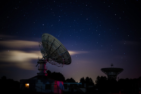 A nighttime scene featuring two large satellite dishes silhouetted against a starry sky. The foreground dish is prominently lit, while a warm glow emanates from a building beneath it. The sky is clear with scattered bright stars, and a few clouds add depth to the composition. Trees line the horizon, adding contrast to the structures.