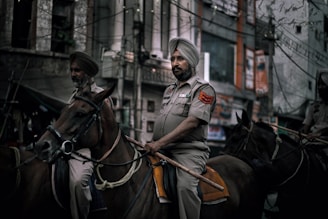 Two uniformed individuals riding horses on a city street, each wearing a turban and appearing to be law enforcement officers. The background shows urban architecture with buildings and signage.