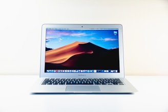 A silver laptop displaying a desert dune wallpaper on its screen, set on a white surface against a plain background. The laptop screen shows a series of application icons at the bottom.