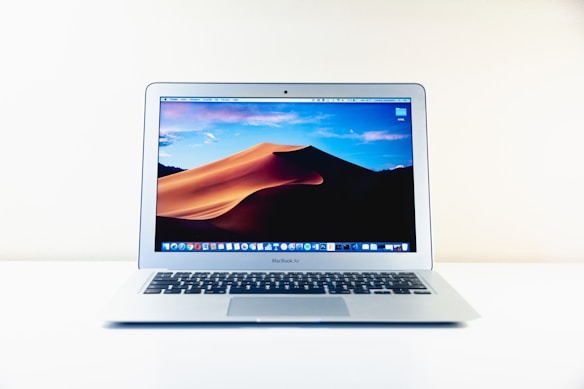 A silver laptop displaying a desert dune wallpaper on its screen, set on a white surface against a plain background. The laptop screen shows a series of application icons at the bottom.