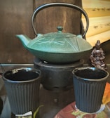 A traditional teapot with intricate engravings is placed on a small stove. Two matching cups are positioned in front, all in a deep green or black hue. In the background, a scroll with Chinese characters and a small statue adds an oriental ambiance.