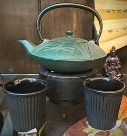 A traditional teapot with intricate engravings is placed on a small stove. Two matching cups are positioned in front, all in a deep green or black hue. In the background, a scroll with Chinese characters and a small statue adds an oriental ambiance.