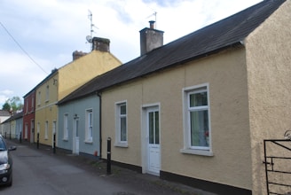 A row of colorful terraced houses lines a narrow street, featuring various shades including yellow, blue, and red. The houses have sloped roofs with chimneys, and some have satellite dishes attached. A car is parked on the road, and there are bollards along the sidewalk.