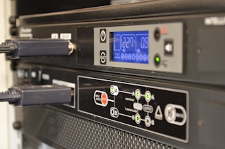 Two electronic devices are stacked, featuring digital displays and several buttons for control. The upper device has a blue screen displaying numbers, with buttons and dials for operation. The lower device showcases a detailed control panel with various symbols and indicators, connected by dark cables.