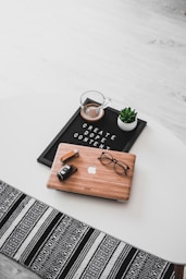 A white oval table with a black felt letter board displaying the phrase 'CREATE DOPE CONTENT'. Next to it, a small potted succulent and a glass cup containing a beverage are placed. A wooden laptop with an Apple logo, a pair of black-rimmed glasses, and a black camera with a lens cap are also arranged neatly. The floor underneath is light wood, and part of a black and white patterned rug is visible at the bottom.