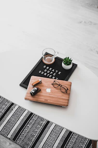 A white oval table with a black felt letter board displaying the phrase 'CREATE DOPE CONTENT'. Next to it, a small potted succulent and a glass cup containing a beverage are placed. A wooden laptop with an Apple logo, a pair of black-rimmed glasses, and a black camera with a lens cap are also arranged neatly. The floor underneath is light wood, and part of a black and white patterned rug is visible at the bottom.