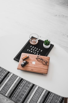 A white oval table with a black felt letter board displaying the phrase 'CREATE DOPE CONTENT'. Next to it, a small potted succulent and a glass cup containing a beverage are placed. A wooden laptop with an Apple logo, a pair of black-rimmed glasses, and a black camera with a lens cap are also arranged neatly. The floor underneath is light wood, and part of a black and white patterned rug is visible at the bottom.