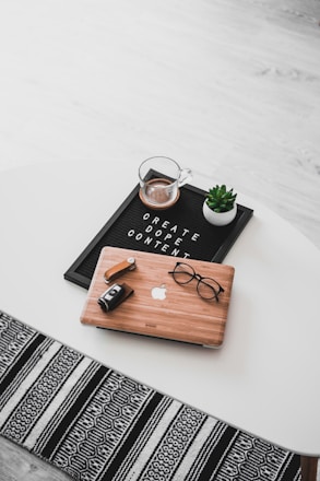 A white oval table with a black felt letter board displaying the phrase 'CREATE DOPE CONTENT'. Next to it, a small potted succulent and a glass cup containing a beverage are placed. A wooden laptop with an Apple logo, a pair of black-rimmed glasses, and a black camera with a lens cap are also arranged neatly. The floor underneath is light wood, and part of a black and white patterned rug is visible at the bottom.