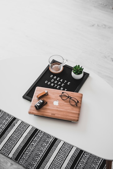 A white oval table with a black felt letter board displaying the phrase 'CREATE DOPE CONTENT'. Next to it, a small potted succulent and a glass cup containing a beverage are placed. A wooden laptop with an Apple logo, a pair of black-rimmed glasses, and a black camera with a lens cap are also arranged neatly. The floor underneath is light wood, and part of a black and white patterned rug is visible at the bottom.
