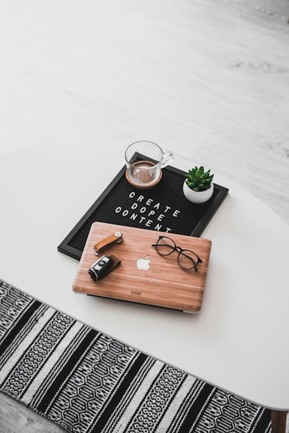 A white oval table with a black felt letter board displaying the phrase 'CREATE DOPE CONTENT'. Next to it, a small potted succulent and a glass cup containing a beverage are placed. A wooden laptop with an Apple logo, a pair of black-rimmed glasses, and a black camera with a lens cap are also arranged neatly. The floor underneath is light wood, and part of a black and white patterned rug is visible at the bottom.