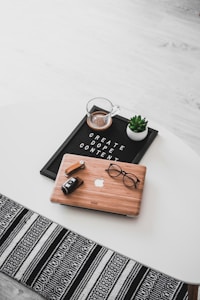 A white oval table with a black felt letter board displaying the phrase 'CREATE DOPE CONTENT'. Next to it, a small potted succulent and a glass cup containing a beverage are placed. A wooden laptop with an Apple logo, a pair of black-rimmed glasses, and a black camera with a lens cap are also arranged neatly. The floor underneath is light wood, and part of a black and white patterned rug is visible at the bottom.