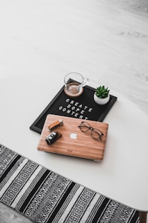 A white oval table with a black felt letter board displaying the phrase 'CREATE DOPE CONTENT'. Next to it, a small potted succulent and a glass cup containing a beverage are placed. A wooden laptop with an Apple logo, a pair of black-rimmed glasses, and a black camera with a lens cap are also arranged neatly. The floor underneath is light wood, and part of a black and white patterned rug is visible at the bottom.