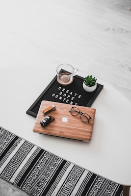 A white oval table with a black felt letter board displaying the phrase 'CREATE DOPE CONTENT'. Next to it, a small potted succulent and a glass cup containing a beverage are placed. A wooden laptop with an Apple logo, a pair of black-rimmed glasses, and a black camera with a lens cap are also arranged neatly. The floor underneath is light wood, and part of a black and white patterned rug is visible at the bottom.