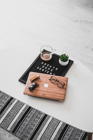 A white oval table with a black felt letter board displaying the phrase 'CREATE DOPE CONTENT'. Next to it, a small potted succulent and a glass cup containing a beverage are placed. A wooden laptop with an Apple logo, a pair of black-rimmed glasses, and a black camera with a lens cap are also arranged neatly. The floor underneath is light wood, and part of a black and white patterned rug is visible at the bottom.