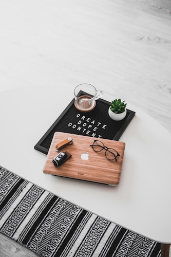 A white oval table with a black felt letter board displaying the phrase 'CREATE DOPE CONTENT'. Next to it, a small potted succulent and a glass cup containing a beverage are placed. A wooden laptop with an Apple logo, a pair of black-rimmed glasses, and a black camera with a lens cap are also arranged neatly. The floor underneath is light wood, and part of a black and white patterned rug is visible at the bottom.