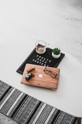 A white oval table with a black felt letter board displaying the phrase 'CREATE DOPE CONTENT'. Next to it, a small potted succulent and a glass cup containing a beverage are placed. A wooden laptop with an Apple logo, a pair of black-rimmed glasses, and a black camera with a lens cap are also arranged neatly. The floor underneath is light wood, and part of a black and white patterned rug is visible at the bottom.