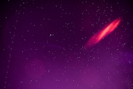 A starry night sky with numerous stars scattered throughout, featuring a prominent, elongated streak of red light in the upper right quadrant. The background is a gradient of deep purples, adding a surreal or cosmic ambiance to the scene.