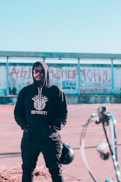 A person wearing a black hoodie with 'Barcelona University' on it is standing in front of a partially visible street art-covered wall. They have their hands in their pockets and are also wearing sunglasses. In the foreground, part of a motorcycle is visible.
