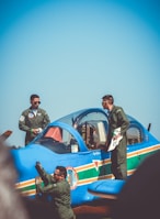 Three people are dressed in military uniforms and are gathered around a bright blue aircraft on a sunny day. Two individuals stand on the wing, while another is adjacent to the plane's side, appearing to inspect or clean it. The aircraft features colorful stripes and insignias on its body.