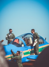Three people are dressed in military uniforms and are gathered around a bright blue aircraft on a sunny day. Two individuals stand on the wing, while another is adjacent to the plane's side, appearing to inspect or clean it. The aircraft features colorful stripes and insignias on its body.