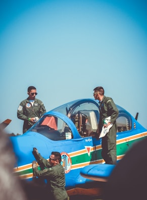Three people are dressed in military uniforms and are gathered around a bright blue aircraft on a sunny day. Two individuals stand on the wing, while another is adjacent to the plane's side, appearing to inspect or clean it. The aircraft features colorful stripes and insignias on its body.