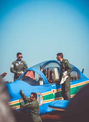 Three people are dressed in military uniforms and are gathered around a bright blue aircraft on a sunny day. Two individuals stand on the wing, while another is adjacent to the plane's side, appearing to inspect or clean it. The aircraft features colorful stripes and insignias on its body.
