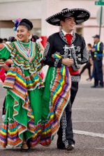 A man and a woman dressed in traditional Mexican costumes are walking side by side, smiling. The woman is wearing a vibrant green dress with colorful stripes and ruffles, while the man is in a black outfit with white embroidery and a large sombrero. An urban background with people in the distance complements the festive atmosphere.