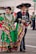 A man and a woman dressed in traditional Mexican costumes are walking side by side, smiling. The woman is wearing a vibrant green dress with colorful stripes and ruffles, while the man is in a black outfit with white embroidery and a large sombrero. An urban background with people in the distance complements the festive atmosphere.
