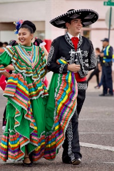 A man and a woman dressed in traditional Mexican costumes are walking side by side, smiling. The woman is wearing a vibrant green dress with colorful stripes and ruffles, while the man is in a black outfit with white embroidery and a large sombrero. An urban background with people in the distance complements the festive atmosphere.