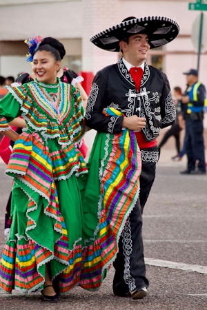 A man and a woman dressed in traditional Mexican costumes are walking side by side, smiling. The woman is wearing a vibrant green dress with colorful stripes and ruffles, while the man is in a black outfit with white embroidery and a large sombrero. An urban background with people in the distance complements the festive atmosphere.