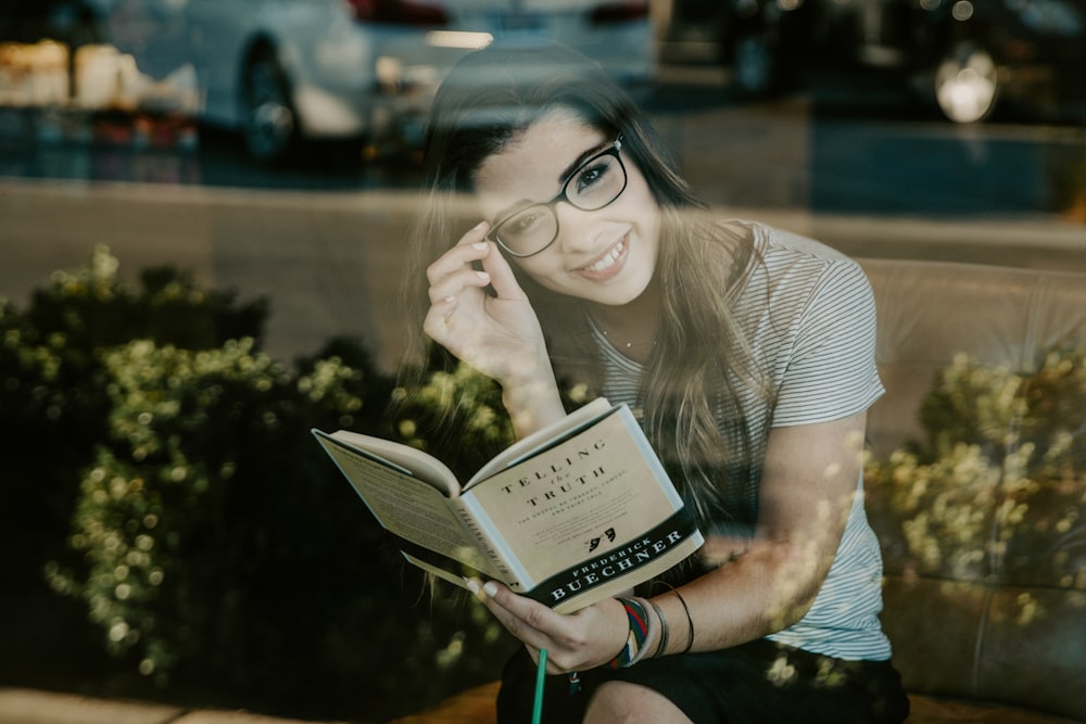 woman smiling wearing black eyeglasses reading book
