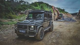 A rugged black off-road vehicle with a custom design is parked on a dirt road, surrounded by trees and mountains. In the background, an excavator is working next to a pile of rocks, suggesting a construction or development site.