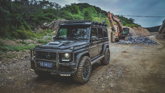 A rugged black off-road vehicle with a custom design is parked on a dirt road, surrounded by trees and mountains. In the background, an excavator is working next to a pile of rocks, suggesting a construction or development site.