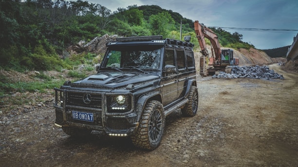 A rugged black off-road vehicle with a custom design is parked on a dirt road, surrounded by trees and mountains. In the background, an excavator is working next to a pile of rocks, suggesting a construction or development site.