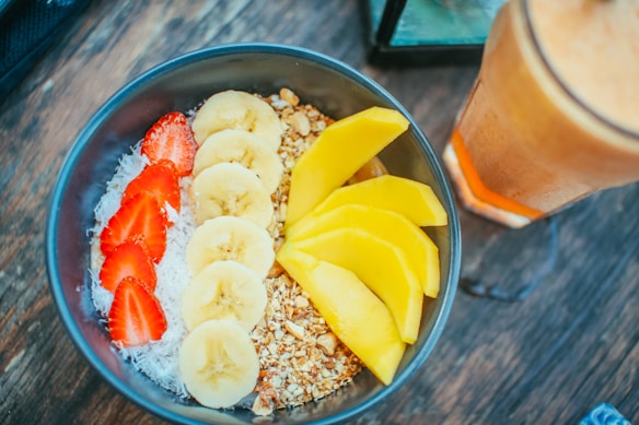 A bowl containing a colorful arrangement of sliced bananas, strawberries, and mangoes, with granola and shredded coconut, sits on a wooden surface. Beside it is a tall glass of a light-brown smoothie or juice.