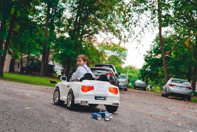 A young child is sitting in a small white toy car on a paved road. The car has a 'Just Married' sign on the back, with tin cans attached and trailing behind. The child is looking back towards the camera, and the setting appears to be a suburban neighborhood with trees and parked cars lining the street.
