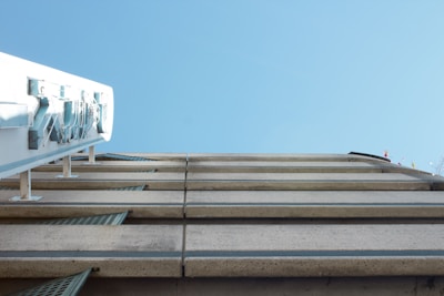 A modern building facade with a clear blue sky. The building has a series of horizontal concrete beams with metal grids, creating a geometric pattern. Equipment is mounted on a metal scaffolding structure to the side.