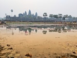 A large historical temple with distinctive spires is reflected in a body of water in the foreground. The scene is populated by numerous visitors walking along the path parallel to the water and exploring the temple grounds. Palm trees dot the landscape, adding to the tropical atmosphere, while pink lotus flowers float on the surface of the water.