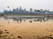 A large historical temple with distinctive spires is reflected in a body of water in the foreground. The scene is populated by numerous visitors walking along the path parallel to the water and exploring the temple grounds. Palm trees dot the landscape, adding to the tropical atmosphere, while pink lotus flowers float on the surface of the water.