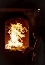 A person stands in front of a furnace or large oven with flames rising inside, casting a warm glow. The individual is holding a piece of paper or cloth, gazing intently at the fire.