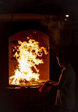 A person stands in front of a furnace or large oven with flames rising inside, casting a warm glow. The individual is holding a piece of paper or cloth, gazing intently at the fire.