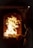 A person stands in front of a furnace or large oven with flames rising inside, casting a warm glow. The individual is holding a piece of paper or cloth, gazing intently at the fire.