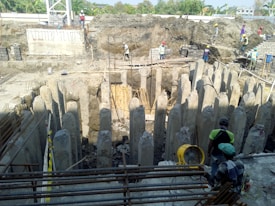 A construction site featuring a circular arrangement of concrete pillars surrounding an excavation area. Several construction workers wearing safety gear, including helmets and vests, are positioned around the site, some on the ground and others on elevated positions. Reinforcement bars and construction equipment are also visible.