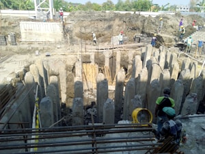 A construction site featuring a circular arrangement of concrete pillars surrounding an excavation area. Several construction workers wearing safety gear, including helmets and vests, are positioned around the site, some on the ground and others on elevated positions. Reinforcement bars and construction equipment are also visible.