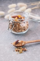 A jar filled with granola, including visible nuts and dried fruits, is placed on a gray table surface. A wooden spoon lies nearby with a serving of granola heaped on it, highlighting the texture and variety of ingredients. Soft-focus background elements include dry, fluffy plant stems.