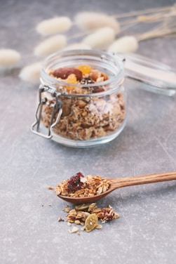 A jar filled with granola, including visible nuts and dried fruits, is placed on a gray table surface. A wooden spoon lies nearby with a serving of granola heaped on it, highlighting the texture and variety of ingredients. Soft-focus background elements include dry, fluffy plant stems.