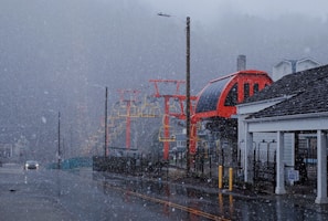 An overcast day with heavy snowfall covering a street, where a red and yellow ski lift structure is visible. The scene includes a car on the wet road, buildings on the sides, and a sign for exit only in the foreground.