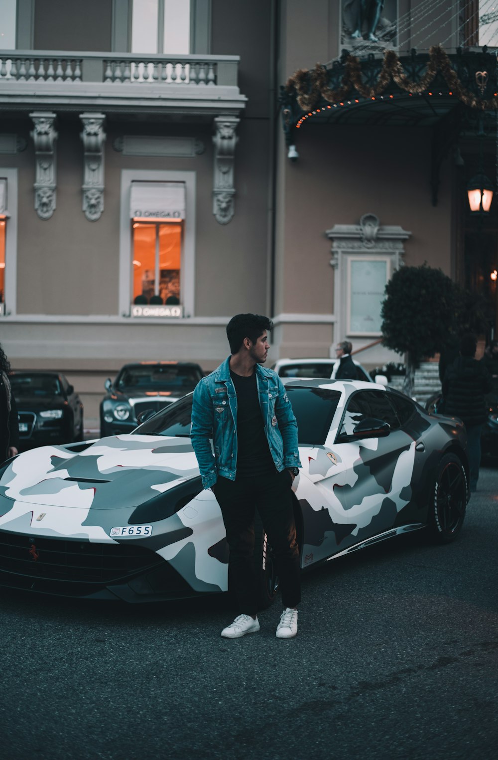 man in blue denim jacket and black pants stands beside parked green and white camouflage sports coupe