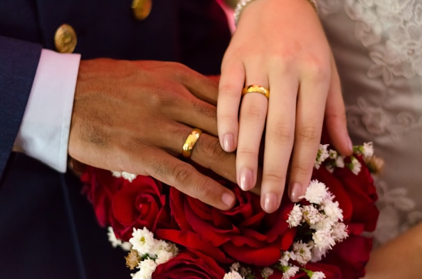 Two hands with wedding rings rest gently on a bouquet of red and white flowers. The hands belong to individuals of different skin tones, symbolizing unity and love. The background features elegant lace fabric, possibly part of a wedding dress.