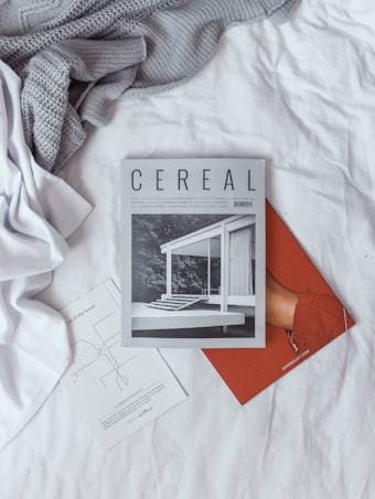 Magazines and papers are laid out on a neatly arranged white bed with a grey knit blanket. The main magazine is titled 'Cereal' and features a monochrome architectural photograph on the cover.